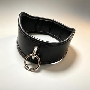 May include: A black collar made of smooth material with stitched edges. The collar features a central silver-toned metal O-ring attached to a smaller metal loop, creating a circular pendant detail on the front of the band.