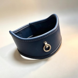 Könnte beinhalten: Ein dunkelblaues Halsband mit glatter, matter Oberfläche und einem mittigen, silberfarbenen Metallring. Das Halsband verfügt über verstärkte, abgesteppte Kanten an Ober- und Unterseite und behält eine geschwungene, strukturierte Form bei.