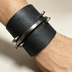 Pode incluir: Uma pulseira larga de couro preto com uma superfície texturizada de grão de seixo. A pulseira é acentuada com uma barra de metal prateado lisa e curva, fixada à banda de couro por dois laços de metal prateado polido em cada lado.