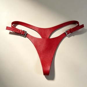 Peut inclure: Un string rouge vif fabriqué dans une matière texturée, doté d'une fine ceinture et d'un panneau central. Le modèle comprend deux sangles latérales réglables avec des boucles en métal argenté, permettant un ajustement personnalisé à la taille.