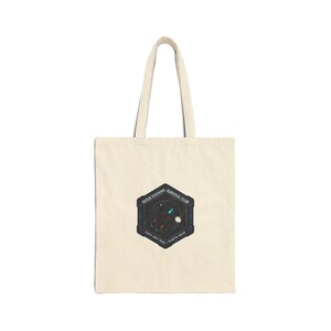 May include: A cream-coloured tote bag featuring a dark blue hexagonal graphic with a space scene. The text reads "Outer Systems Survival Club" above and "Plan A: Don't Panic. Plan B: Math" below the graphic.