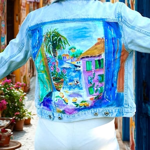 Peut inclure: Veste en jean bleu clair avec une scène côtière peinte à la main sur le dos. L'œuvre d'art représente un palmier, des bâtiments, des bateaux et des accents colorés. La veste a un design classique avec des boutons.
