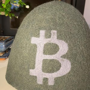Könnte beinhalten: Ein salbeigrüner Filzhut mit einem großen weißen Bitcoin-Symbol auf der Vorderseite. Der Hut hat eine abgerundete, kuppelförmige Form und ein weiches, strukturiertes Aussehen. Das Bitcoin-Symbol ist deutlich sichtbar.