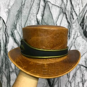 May include: A tan top hat with a textured crocodile skin pattern. The hat features a wide black band around the base, accented by a thin, metallic green trim that encircles the crown. The brim is slightly curved, and the hat has a structured shape.