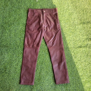 Könnte beinhalten: Eine bordeauxfarbene Lederhose mit klassischem Fünf-Taschen-Design. Die Hose hat einen Knopf- und Reißverschluss, Gürtelschlaufen und verstärkte Knieeinsätze. Die Hose wird auf einer grünen Oberfläche präsentiert.