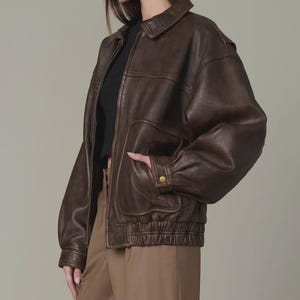 May include: A dark brown leather bomber jacket featuring a classic collar, a front zipper closure, and long sleeves with buttoned cuffs. The jacket includes two large front patch pockets and an elasticized hemline, providing a structured fit.