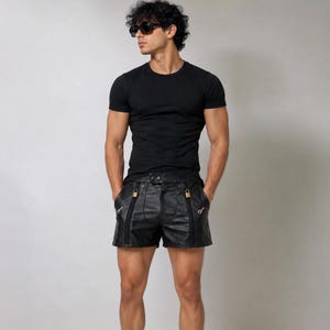 Könnte beinhalten: Ein schwarzes kurzärmeliges T-Shirt mit Rundhalsausschnitt kombiniert mit einer schwarzen Ledershorts. Die Shorts verfügen über zwei auffällige vertikale Reißverschlusstaschen auf der Vorderseite, die mit kleinen Metallschlössern verziert sind.