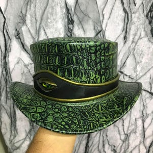 Può includere: Un cappello a cilindro con una texture in pelle di coccodrillo verde e nera. Il cappello presenta un'ampia fascia nera attorno alla base con un bordo color oro e un prominente occhio di rettile verde realistico incastonato al centro.