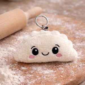 Puede incluir: Un llavero de peluche blanco con forma de dumpling con una cara sonriente, ojos negros y mejillas rosadas. El llavero tiene un anillo plateado y está colocado sobre una tabla de cortar de madera espolvoreada con harina.