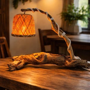 Puede incluir: Lámpara de mesa rústica con base de madera natural y una rama curvada que sostiene una pantalla tejida naranja. La rama está envuelta con una cuerda blanca. La lámpara está sobre una mesa de madera.