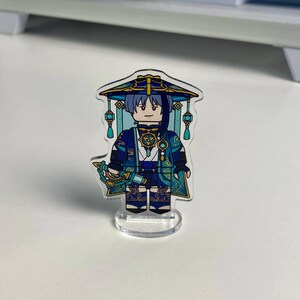 K&ouml;nnte beinhalten: Eine transparente Acrylfigur eines Charakters in einem blau-goldenen Outfit mit passendem Hut. Die Figur steht auf einem transparenten Sockel. Der Charakter hat einen strengen Ausdruck und h&auml;lt eine Waffe.