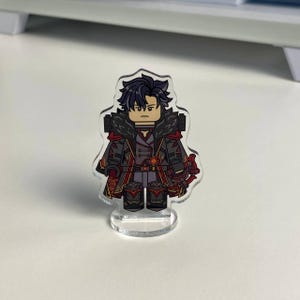 K&ouml;nnte beinhalten: Klare Acrylfigur eines Charakters mit dunklem Haar und ernstem Gesichtsausdruck. Die Figur tr&auml;gt ein detailliertes Outfit mit roten und schwarzen Akzenten und steht auf einem klaren Sockel. Das Design &auml;hnelt einer Miniaturfigur.