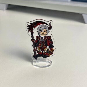 K&ouml;nnte beinhalten: Eine transparente Acrylfigur eines Charakters mit wei&szlig;en Haaren, H&ouml;rnern und einem rot-schwarzen Outfit, der eine Sense h&auml;lt. Die Figur steht auf einem transparenten Sockel.