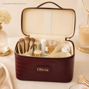 May include: A deep burgundy makeup train case with a crocodile-embossed texture and a gold zip. The front features a rectangular patch with the name Olivia in gold lettering. The interior is lined in cream fabric with dividers and elastic loops.