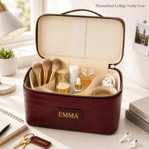 May include: A deep burgundy vanity case with a crocodile-embossed texture and a gold zip. The front features a rectangular patch with the name EMMA in gold lettering. The interior is lined in cream fabric with compartments for brushes.