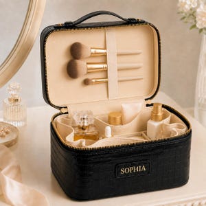 May include: A black rectangular makeup case with a crocodile-embossed texture and a top handle. The front features a gold-lettered name tag reading SOPHIA. The interior is lined in beige fabric, containing three makeup brushes and various bottles.