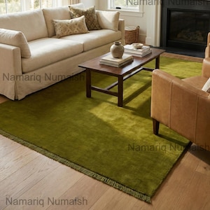 Peut inclure: Un salon avec un grand tapis vert olive à bordure frangée. Le tapis est sous une table basse en bois foncé et un canapé crème avec des coussins décoratifs. Un fauteuil en cuir est à droite. Le tapis mesure environ 2,4 m x 3 m.