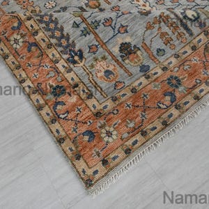 May include: A portion of a hand-knotted rug with a detailed floral pattern. The rug features a light blue field with an orange border. The border has a repeating pattern of floral and geometric elements. The rug is partially rolled up, revealing the underside.