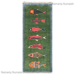 Könnte beinhalten: Ein rechteckiger grüner Teppich mit einem sich wiederholenden Muster aus bunten Fischen. Die Fische sind in Rot-, Orange- und Schwarztönen gehalten und weisen aufwendige Designs auf. Der Teppich hat einen blauen Rand und Fransen.
