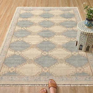 May include: A rectangular rug with a diamond pattern in light blue and beige. The rug has a fringed border and is on a wooden floor. A small side table with a floral design and a plant in a blue pot are next to the rug.