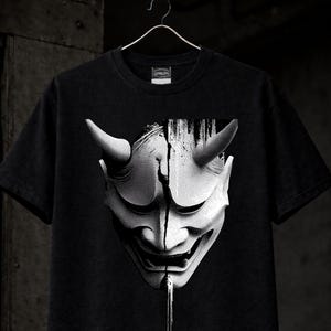 May include: Black t-shirt featuring a detailed, grayscale illustration of a Japanese demon mask. The mask has horns and a stern expression, with a vertical paint-like effect down the center. The shirt is hanging on a hanger.