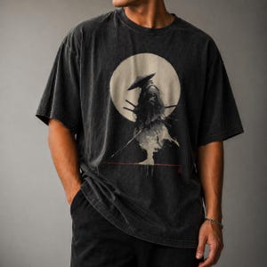 May include: Dark gray t-shirt featuring a samurai warrior illustration. The design shows a samurai in front of a full moon. The shirt has a vintage, washed look and a relaxed fit.