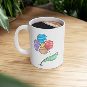 May include: White ceramic coffee mug with a colorful, hand-drawn flower design. The flower has blue, purple, red, brown, and yellow petals, with a green stem and leaf. The mug is filled with dark coffee.
