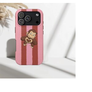 Könnte beinhalten: Eine pink-rot gestreifte Handyhülle mit einem Cartoon-Affen, der mit seinen Händen ein Herz formt. Die Hülle hat einen erhöhten Rand um die Kameralinsen und eine glatte Oberfläche.