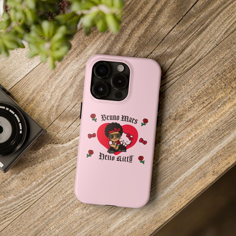 Puede incluir: Funda de tel&eacute;fono rosa con un gr&aacute;fico de Bruno Mars y Hello Kitty dentro de un coraz&oacute;n rojo. El dise&ntilde;o incluye rosas y el texto "Bruno Mars" y "Hello Kitty". La funda est&aacute; sobre una superficie de madera.