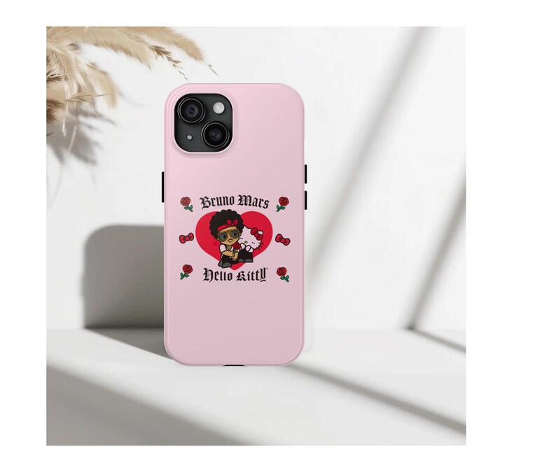 Puede incluir: Funda de iPhone rosa claro con un gr&aacute;fico de Bruno Mars y Hello Kitty en un coraz&oacute;n rojo. La funda tiene botones laterales negros y el texto "Bruno Mars" y "Hello Kitty". Tambi&eacute;n se presentan rosas y lazos rojos.