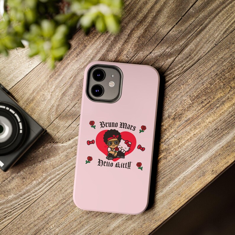 Puede incluir: Funda de tel&eacute;fono rosa con un dibujo animado de Bruno Mars y Hello Kitty dentro de un coraz&oacute;n rojo. La funda tiene el texto "Bruno Mars" y "Hello Kitty" en una fuente decorativa, con rosas y lazos. La funda del tel&eacute;fono est&aacute; sobre una superficie de madera.