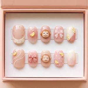 May include: A set of ten artificial nails in a pink box. The nails feature various designs, including a monkey face, a bow, a banana, and polka dots. Some nails have a French tip design with white polka dots.
