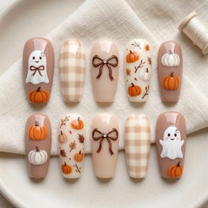 May include: A set of ten press-on nails featuring an autumnal theme with beige, brown, and white colours. Designs include 3D white ghosts with crystal accents, brown bows, orange and white pumpkins, and a beige checkered pattern on a glossy finish.