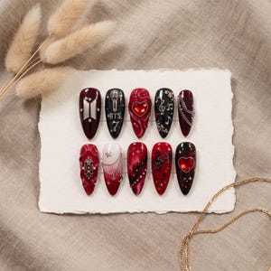 May include: A set of ten almond-shaped press-on nails featuring a dark red and black color palette. Designs include a silver BTS logo, a microphone with the text BTS 7, red heart gems, musical notes, silver chains, stars, and ornate filigree.
