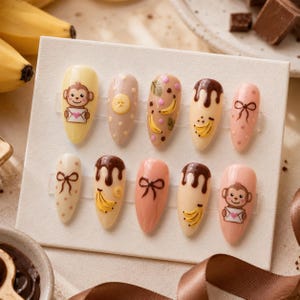 Op de afbeelding: Een set van tien amandelvormige kunstnagels met diverse ontwerpen, waaronder getekende aapjes met enveloppen, bruine strikjes, gele bananenillustraties, bruine chocoladedruppels en stippen in crème- en roze tinten.