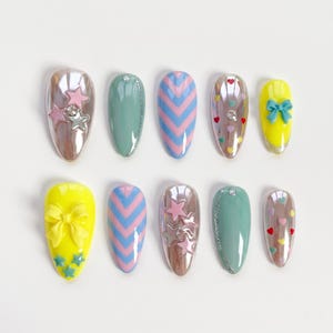 May include: A collection of ten oval-shaped artificial nails. Designs include pink and silver stars, blue and pink chevron stripes, and yellow nails with bows and stars. Some nails have heart and glitter accents.
