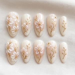 May include: A set of ten almond-shaped artificial nails. Each nail is painted a pale pink with a white floral design and gold accents. The flowers are three-dimensional, adding texture to the design. The nails are arranged on a white surface.