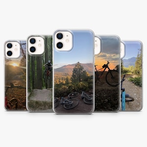 May include: A collection of five iPhone cases featuring scenic mountain biking images. The cases showcase various outdoor scenes, including a rider in mid-air, bicycles on a trail, and mountain landscapes at sunset. The cases have a clear, protective design.