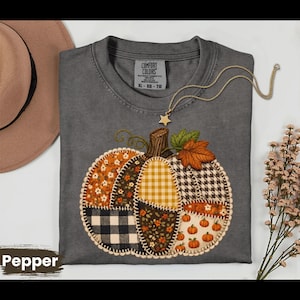 May include: A charcoal gray short-sleeve t-shirt featuring a large pumpkin graphic composed of various fabric-style patches including plaid, gingham, and floral patterns. The shirt has a Comfort Colors label and a gold-tone star pendant necklace.