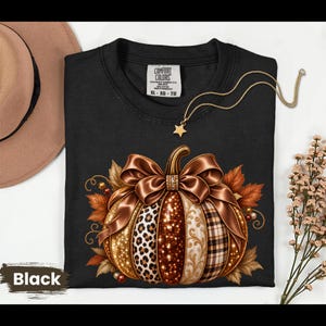 May include: A black t-shirt featuring a graphic of a pumpkin adorned with a large brown satin bow. The pumpkin segments display varied textures including gold glitter, leopard print, and plaid patterns, surrounded by autumn leaves and berries.