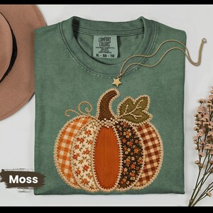 May include: A moss green t-shirt featuring a large pumpkin graphic composed of various fabric patterns including orange gingham, floral prints, and solid corduroy. The shirt has a Comfort Colors label at the neck and a gold star pendant necklace.