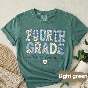 Puede incluir: Una camiseta de manga corta color verde salvia con el texto FOURTH GRADE en fuente de bloque con patrones de cuadros vichy azules y flores blancas. Debajo del texto, el nombre Miss Emma está escrito en cursiva, con un gráfico de margarita.