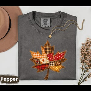 May include: A charcoal gray crewneck t-shirt featuring a large, central maple leaf graphic composed of various fabric patches in plaid, polka dot, and floral patterns. The shirt includes a Comfort Colors label and a gold-tone star pendant necklace.