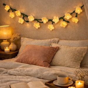 May include: A cozy bedroom scene featuring a bed with soft pillows and a plush comforter. A garland of crochet flowers with lights hangs above the bed. A tray with a cup of coffee, candles, and a book sits on the bed, creating a warm and inviting atmosphere.