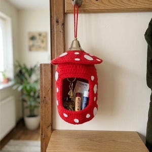May include: A red and white crocheted mushroom-shaped wall organizer hanging from a metal hook. The hollow center contains a set of keys, a ticket, and two lip balm tubes. The top features a metallic bell-shaped cap with a red hanging cord.