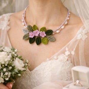 May include: A crochet necklace featuring three small light pink flowers and several dark green and sage green crocheted leaves. The necklace is strung on a double-strand cord accented with small white pearls and tiny metallic beads.