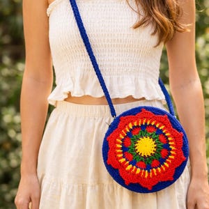 May include: A round, crocheted crossbody bag featuring a vibrant, multi-colored mandala pattern in shades of bright blue, red, green, and yellow. The bag has a long, matching blue crocheted strap and a circular main compartment with a textured finish.