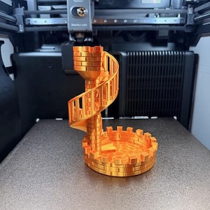 May include: A 3D-printed orange castle tower with a spiral staircase and crenelated walls. The model is on a 3D printer bed, showcasing the manufacturing process. The base of the tower is circular, and the staircase winds upwards.