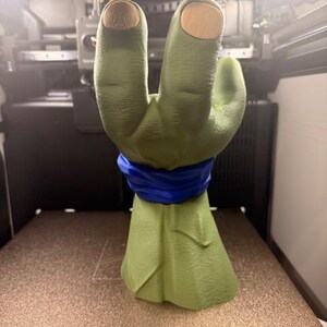 K&ouml;nnte beinhalten: Eine gr&uuml;ne, 3D-gedruckte Handskulptur mit einem blauen Band um das Handgelenk. Die Fingerspitzen sind hellbraun. Die Skulptur befindet sich auf einer braunen Oberfl&auml;che in einem 3D-Drucker.