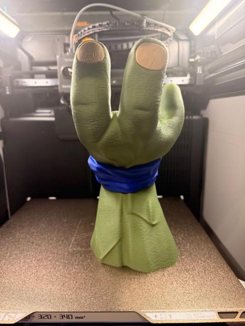 K&ouml;nnte beinhalten: Eine gr&uuml;ne, 3D-gedruckte Handskulptur mit einem blauen Band um das Handgelenk. Die Fingerspitzen sind hellbraun. Die Skulptur befindet sich auf einer braunen Oberfl&auml;che in einem 3D-Drucker.